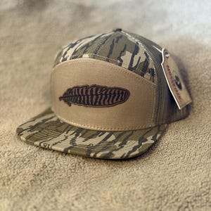 Puede incluir: Una gorra de camionero de camuflaje marr&oacute;n y verde con un parche de cuero marr&oacute;n que presenta un dise&ntilde;o de pluma marr&oacute;n y negro. La gorra tiene una parte trasera de malla y una etiqueta negra que dice "PROSPER OUTDOORS".