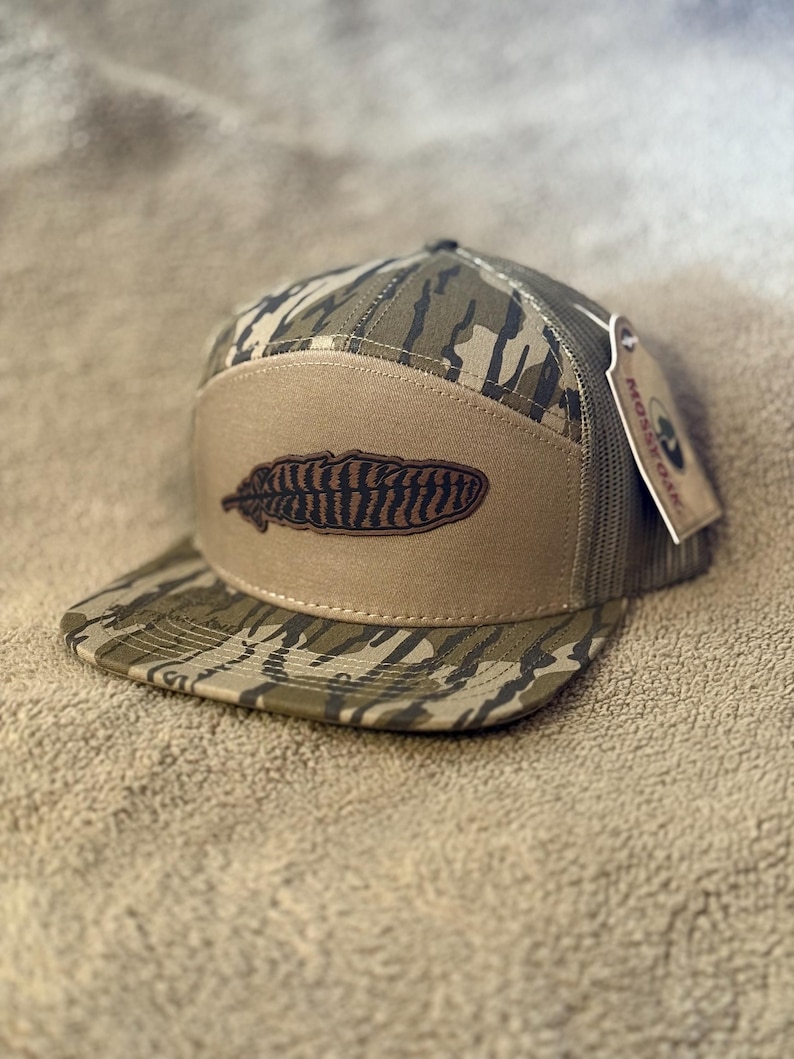 Puede incluir: Una gorra de camionero de camuflaje marr&oacute;n y verde con un parche de cuero marr&oacute;n que presenta un dise&ntilde;o de pluma marr&oacute;n y negro. La gorra tiene una parte trasera de malla y una etiqueta negra que dice "PROSPER OUTDOORS".