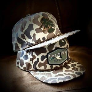 May include: Two camouflage baseball caps stacked on top of each other. The caps have a brown and white pattern. One cap has a patch that says "HAT DOWN" with a duck graphic. The other cap has a duck graphic and the letters "HDW".