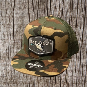 May include: A camouflage trucker hat with a black patch that says "Hat Down Waterfowl". The hat has a black and white logo for "Deckys Classics Original" on the side.