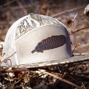 Puede incluir: Una gorra de b&eacute;isbol de camuflaje gris y marr&oacute;n con un parche de cuero marr&oacute;n en forma de pluma en la parte delantera.