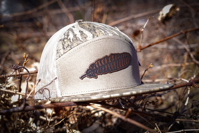 Puede incluir: Una gorra de b&eacute;isbol de camuflaje gris y marr&oacute;n con un parche de cuero marr&oacute;n en forma de pluma en la parte delantera.