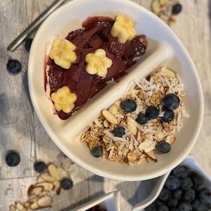 May include: A white bowl divided into two sections. One section contains a red smoothie topped with yellow star-shaped banana slices. The other section contains granola, shredded coconut, almonds, and blueberries.