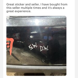 May include: A white vinyl decal applied to a black surface featuring the letters S.W. and D.W. in a stylized, jagged font. To the left, a portion of a red graphic design is visible, showing a curved, textured shape against the dark background.