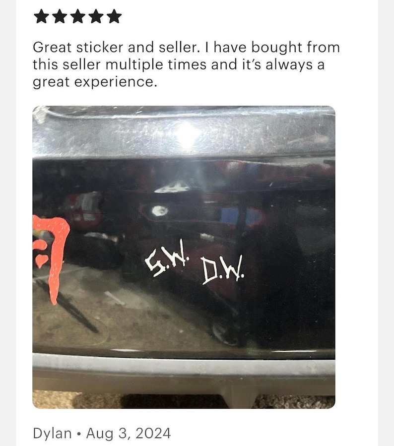 May include: A white vinyl decal applied to a black surface featuring the letters S.W. and D.W. in a stylized, jagged font. To the left, a portion of a red graphic design is visible, showing a curved, textured shape against the dark background.