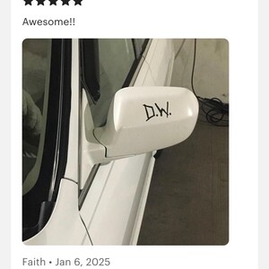 May include: A white car side mirror featuring a black decal with the letters D.W. in a stylized font. The mirror housing is smooth and curved, attached to the white body of the vehicle near the window frame, showing a clean and simple exterior design.