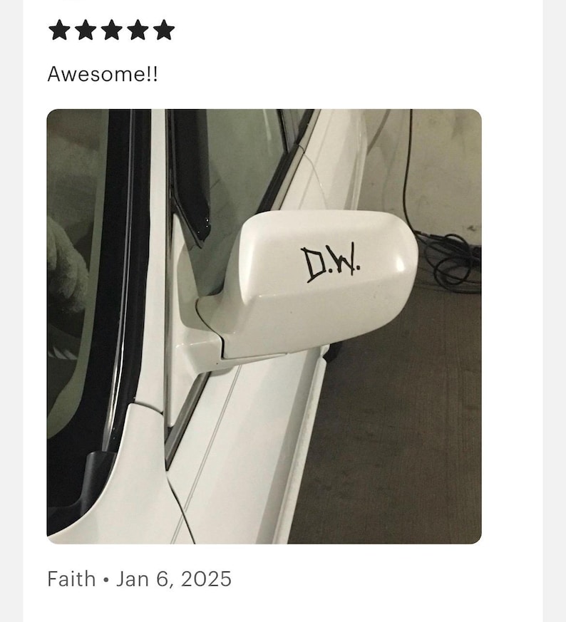May include: A white car side mirror featuring a black decal with the letters D.W. in a stylized font. The mirror housing is smooth and curved, attached to the white body of the vehicle near the window frame, showing a clean and simple exterior design.