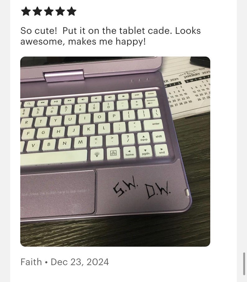 May include: A lavender tablet keyboard case featuring a white QWERTY keyboard layout and a trackpad area. The trackpad surface is marked with the black handwritten initials S.W. and D.W. The device is positioned next to a January 2025 calendar page.