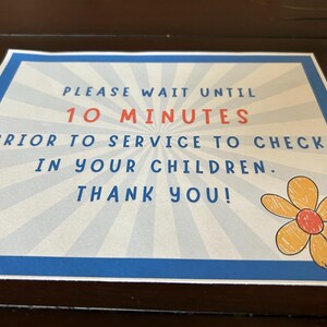 May include: A blue and white sign with a yellow flower graphic. The sign reads "Please wait until 10 minutes prior to service to check in your children. Thank you!"