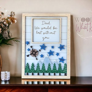 May include: A framed wooden sign with the text "Dad. We would be lost without you." Features a compass, blue stars, and mountain and tree cutouts. The sign has a light blue background and a cream-colored frame.