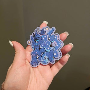 Op de afbeelding: Een blauwe en paarse glitter sticker met een cartoon pony met een kroon, vleugels en een halve maan. De pony heeft blauwe ogen en een roze neus. De sticker is omringd door sterren.