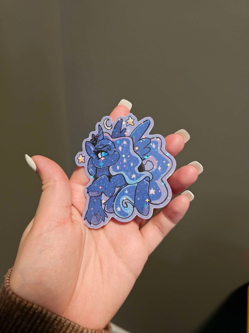 Op de afbeelding: Een blauwe en paarse glitter sticker met een cartoon pony met een kroon, vleugels en een halve maan. De pony heeft blauwe ogen en een roze neus. De sticker is omringd door sterren.