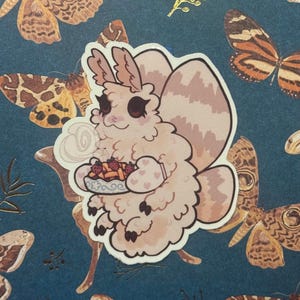 May include: A sticker featuring a cartoon moth character holding a plate of food with strawberries. The moth is light pink with a fluffy appearance. The background is a dark teal color with butterfly and moth illustrations.