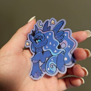 Op de afbeelding: Een blauwe en paarse cartoon pony met een halve maan en sterren op zijn manen. De pony heeft een kroon en blauwe ogen. De sticker is op een witte achtergrond.