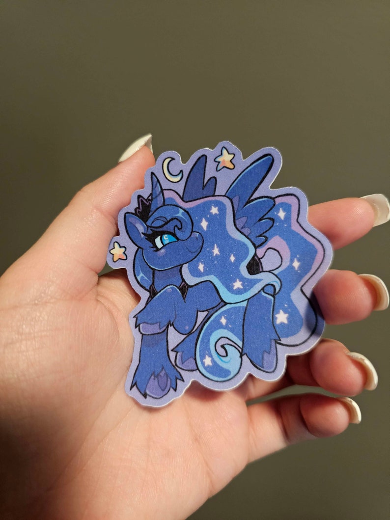 Op de afbeelding: Een blauwe en paarse cartoon pony met een halve maan en sterren op zijn manen. De pony heeft een kroon en blauwe ogen. De sticker is op een witte achtergrond.