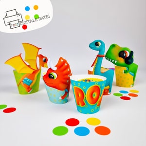 Könnte beinhalten: Vier bunte Cupcake-Verpackungen mit Dinosaurier-Motiven. Die Verpackungen sind blau, grün, gelb und orange mit weißen Punkten. Eine Verpackung hat das Wort "Roar" in orangefarbenen Buchstaben.