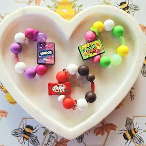 May include: Three beaded bracelets with candy-themed charms, including a "Nerds" box, a "Sour Patch" candy box, and a "Kit Kat" bar. The bracelets feature colourful beads in various sizes and colours. The bracelets are displayed in a white heart-shaped dish.
