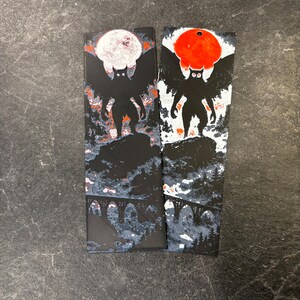 Puede incluir: Dos marcadores negros con un diseño de criatura. Cada marcador presenta una silueta de una figura similar a un murciélago con una luna llena o un sol rojo en el fondo. La parte inferior de los marcadores muestra un puente.