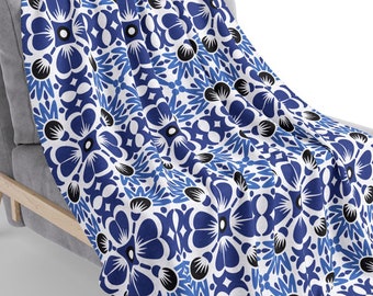 May include: A blue and white floral patterned throw blanket draped over a gray armchair. The blanket features a repeating pattern of stylized flowers with black centers.