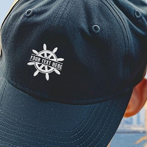 May include: Two black baseball caps with white embroidered nautical wheel designs and the text "Your Text Here".