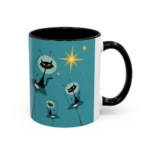 May include: A white ceramic mug with a black rim and handle. The mug features a retro-style design with three black cats in spaceships flying through a blue sky with yellow stars.