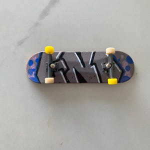 Pode incluir: Um pequeno skate cinza com desenhos abstratos em preto e branco, detalhes azuis e rodas amarelas.