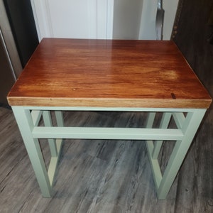 May include: A wooden table top with a light brown wood grain finish sits on a light green metal frame with a single shelf. The table is on a grey floor.
