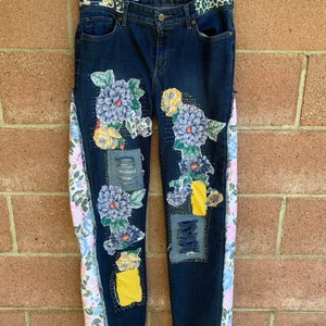 May include: Dark wash denim jeans with floral and patchwork details. The jeans feature blue and yellow floral appliques, distressed patches, and patterned side panels. The waistband has a leopard print design. The brand name "Eddie Bauer" is visible.