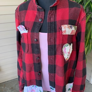 May include: Red and black plaid flannel shirt with floral patches and a ruffled hem. The shirt has a pink undershirt and is displayed on a black mannequin. The shirt has a unique, handcrafted appearance.