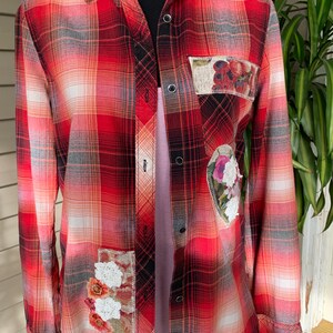 May include: Red plaid flannel shirt with floral patches. The long-sleeved shirt features a classic plaid pattern in shades of red, white, and gray. Decorative patches with embroidered flowers and crochet details add a unique touch to the garment.
