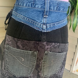 May include: A denim skirt with a patchwork design. The skirt features a blue denim waistband, a black denim midsection, and a patterned gray lower section with two back pockets. The skirt is a unique, handmade garment.