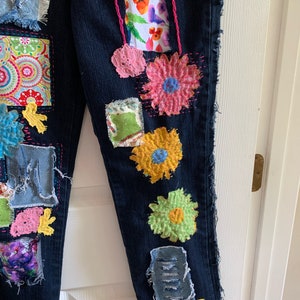 May include: Dark blue denim jeans with colorful fabric patches and embroidered flower details. Patches include floral prints, textured flowers, and frayed denim accents. The jeans have a unique, handcrafted appearance.