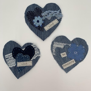 May include: Three denim heart-shaped patches with decorative elements. Each heart features a different design, including lace, fabric flowers, and the words "Love" and "Be Amazing" on small labels. The hearts are arranged on a white background.