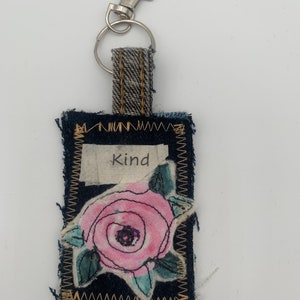 May include: A handmade denim keychain with a silver clasp. The rectangular tag features a pink rose applique and the word "Kind" on a white fabric patch. The edges are stitched with a contrasting thread.