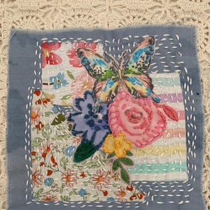 May include: A textile art piece featuring a butterfly and floral design. The butterfly has blue, yellow, and green wings. The flowers are pink, blue, and yellow. The artwork is on a square of blue fabric with white stitching.