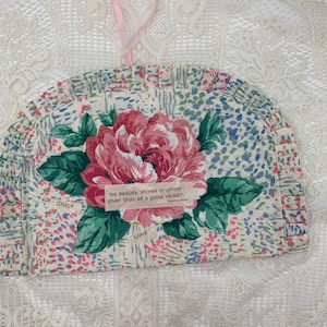 May include: A floral tea cozy with a large pink peony and green leaves. The fabric has a small floral pattern in pink, blue, and green. A pink ribbon hangs from the top. Text on the cozy reads "No beauty shines brighter than that of a good HEART."