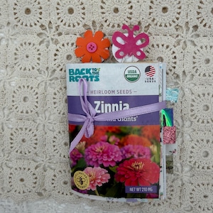 May include: A packet of Zinnia 'and Giants' heirloom seeds, tied with a purple ribbon. The packet features a colorful image of the flowers and the text "Back to the Roots". Adorned with orange flower and pink butterfly embellishments.