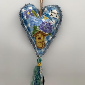 May include: A heart-shaped fabric ornament with a blue gingham pattern and denim edges. It features embroidered flowers, a birdhouse, and a tassel. The word "Dream" is embroidered on a small tag.