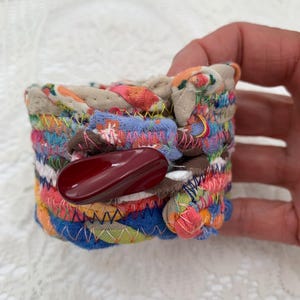 May include: A colorful fabric bracelet with a large, dark red button. The bracelet is made of various fabric scraps in shades of blue, pink, orange, and green, stitched together with contrasting thread. The button is oval-shaped.