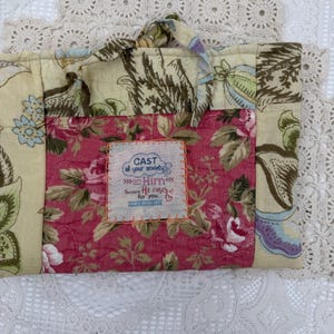 May include: A quilted fabric tote bag with a floral pattern in pink, green, and cream. The bag features a handle and a patch with the text "CAST all your anxiety on Him because He cares for you."