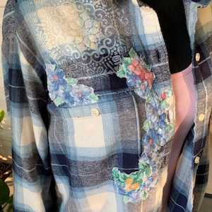 May include: Blue plaid flannel shirt with floral embroidery. The shirt features a classic plaid pattern in shades of blue and white, with embroidered floral accents on the front and pocket. The shirt has a button-down front and a single chest pocket.