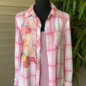 May include: Pink and white plaid button-down shirt with floral embroidery on the front. The shirt has a collared neckline, long sleeves, and a pocket on the chest. A pink tank top is worn underneath.