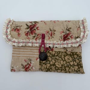 May include: A quilted patchwork clutch with a floral pattern in shades of red, cream, and green. The flap is trimmed with ruffled fabric and a decorative ribbon. A large, dark button secures the closure.
