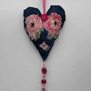 May include: A denim heart-shaped ornament with pink floral fabric appliqués. A pink button and ribbon are attached at the top for hanging. A beaded strand of pink and red beads dangles from the bottom.