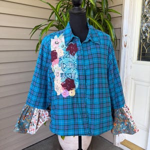 May include: A teal plaid shirt with floral embellishments and bell sleeves. The shirt features a vertical panel of embroidered flowers and lace. The bell sleeves have a floral pattern. The shirt is displayed on a black mannequin.
