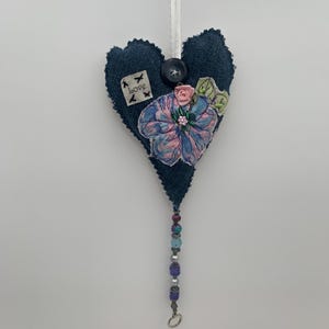 May include: A dark blue fabric heart-shaped ornament with a floral design and beaded tassel. The heart has a button and a small tag that says "love". The flower is pink and blue with green leaves.