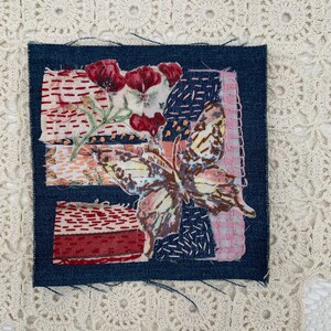 May include: A square textile art piece with a dark blue denim border. The artwork features a butterfly and floral design with red, pink, and orange hues. The background is a cream-colored, crocheted doily.
