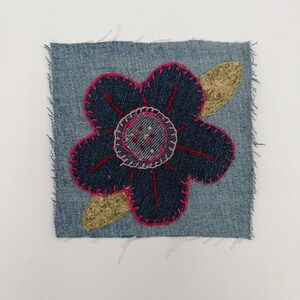 May include: A denim patch featuring a dark blue flower with pink stitching and a light pink center. The flower is surrounded by a pink outline and has a gold-colored leaf. The edges of the denim are frayed.