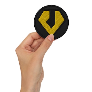 Puede incluir: Un parche bordado negro y amarillo con forma de diamante y una letra V en su interior.