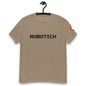 Könnte beinhalten: Ein beiges T-Shirt mit einem schwarzen "ROBOTECH"-Logo auf der Vorderseite. Das Shirt hat einen roten und weißen Aufnäher auf dem rechten Ärmel.